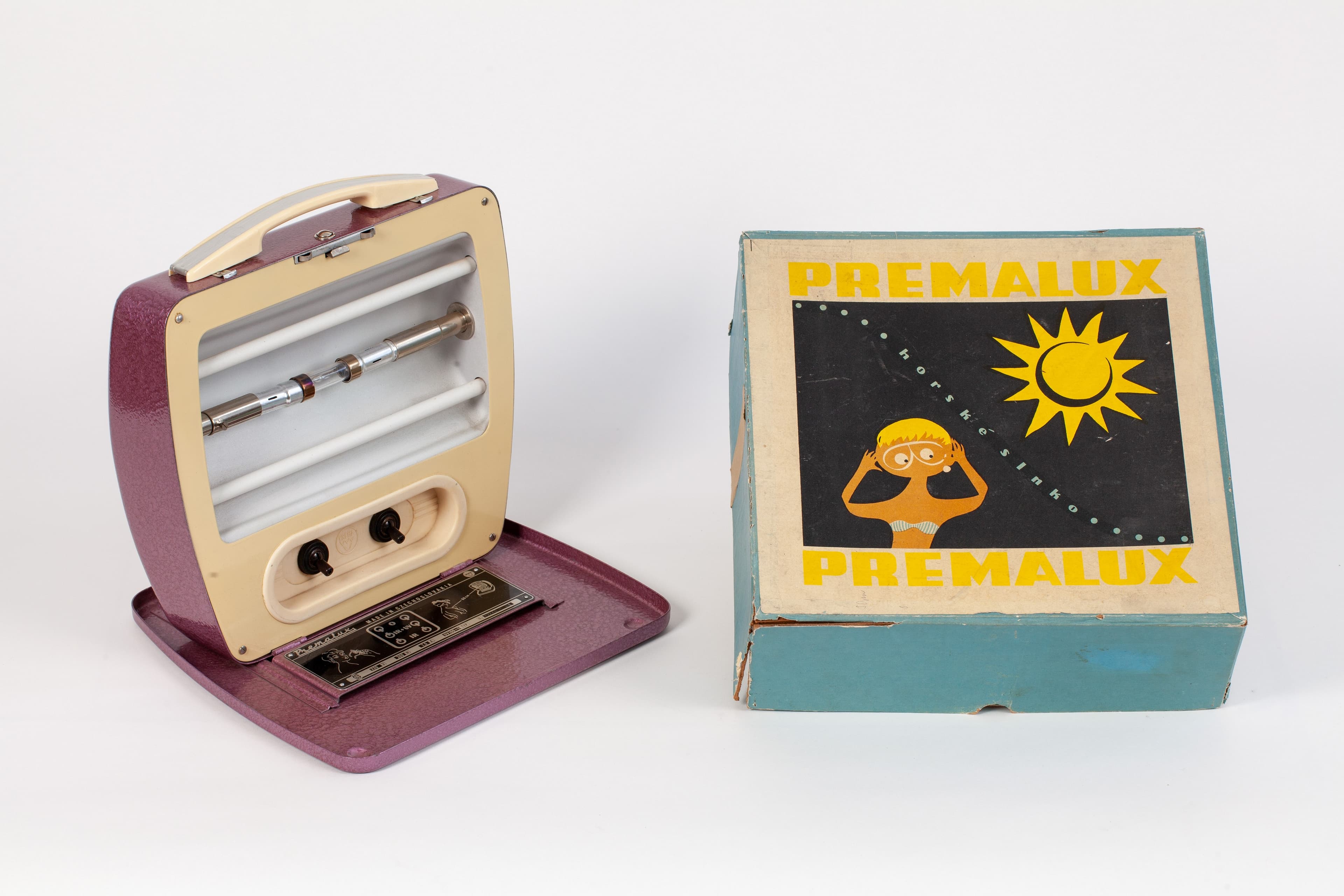 A portable infrared lamp on a stand, displayed together with its original Premalux box. The lamp has a purple and beige casing with a metal reflector at the front.